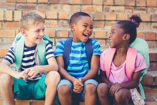 Three school children sitting together on steps, laughing, wearing backpacks